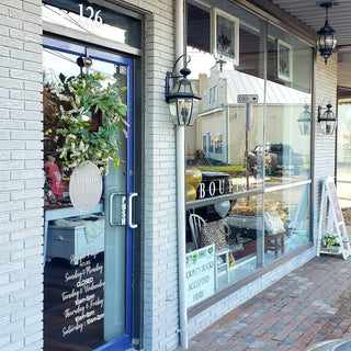 Exterior of boutique storefront with large display windows, hanging wreath on the door, and visible shop hours sign.