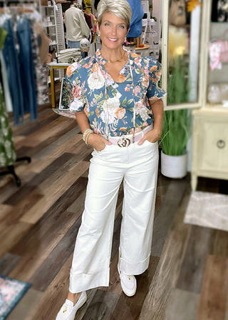 Blue floral puff sleeve blouse paired with white wide-leg pants, pink belt, and white loafers in a boutique setting.