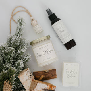 Hollowood Fresh Cut Balsam candle, spray bottle, and soap on a white surface with a small tree and wood logs.