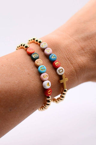 Three beaded bracelets stacked on a person's wrist. The bracelets feature colorful letter beads spelling out 'JESUS' and 'LOVE' with a gold cross charm, accented by gold-toned spacer beads on a light gray background.