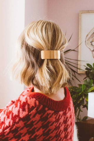 A person shown from the back wearing a wide, rectangular metallic gold hair clip to hold up a blonde bob, in an indoor setting with a pink wall and plants in the background.