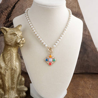 A pearl beaded necklace with a gold-tone cross-shaped pendant featuring colorful inlay stones, displayed on a white jewelry bust next to a gold cat figurine.