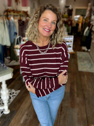 A smiling woman with blonde curly hair stands in a clothing boutique wearing a burgundy and white striped long-sleeve knit sweater, light blue jeans, and a chunky silver chain necklace.