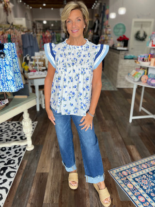 A woman modeling a white smocked blouse with blue floral print and blue ruffled sleeves, paired with blue jeans and beige slide sandals, inside a boutique store.