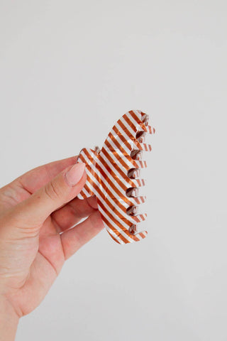 A hand holding a claw hair clip decorated with diagonal orange and white stripes, set against a plain light gray background.