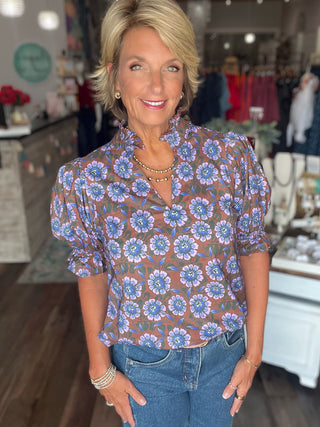 A woman with blonde hair wearing a brown sheer blouse with a blue and purple floral pattern, paired with blue jeans, standing in a clothing boutique.