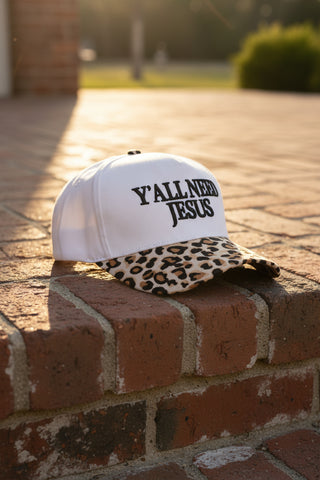 White cap with leopard print brim and 'Y'all Need Jesus' text on a brick step in the sunshine