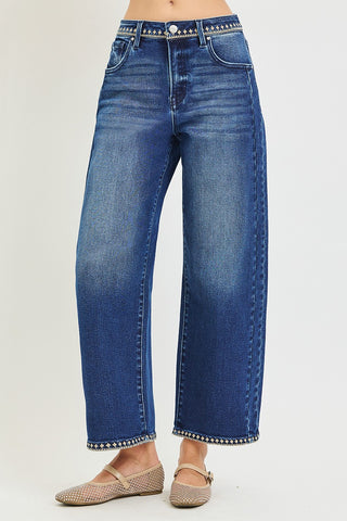 A person wearing blue wide-leg cropped jeans with silver stud detailing along the waistband and hem, paired with beige flat shoes, shown against a plain light gray studio background.
