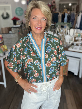 A woman with blonde hair stands in a boutique wearing a dark green short-sleeve button-up blouse with a colorful floral pattern, white pants, and gold jewelry.