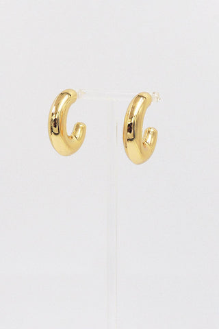 A pair of polished gold huggie hoop earrings displayed on a clear stand against a plain white background.