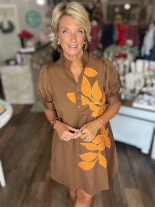 A woman modeling a brown short-sleeve tunic dress with orange leaf embroidery, standing inside a clothing boutique.