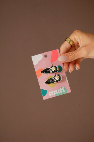 A hand holding a pink card displaying a pair of black and white kitten-themed stud earrings against a solid brown background.