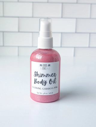 A bottle of shimmer body oil in glowing goddess pink, featuring a white spray top and a white label with black text, set against a white subway tile background.