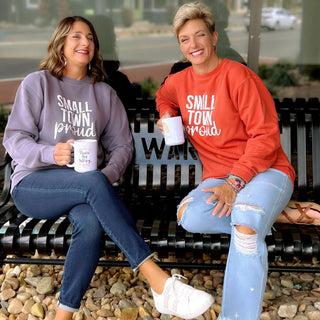 Two women sitting on a bench wearing Small Town Proud sweatshirts, holding coffee mugs.