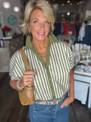 A woman wearing a green and white vertically striped short-sleeve button-up shirt, blue jeans, and a tan shoulder bag, standing inside a boutique.