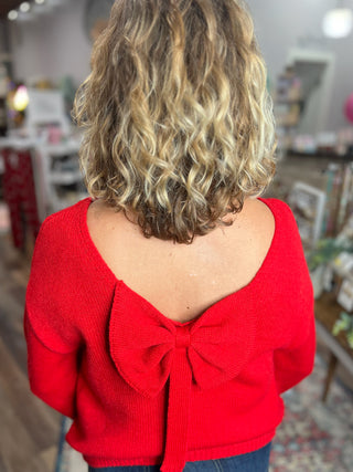 Person wearing a red sweater with a bow detail on the back, standing in an indoor setting.