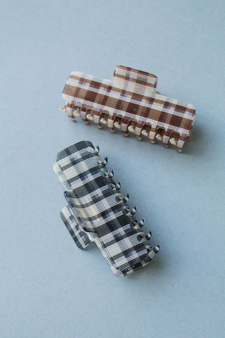 Two fabric hair claw clips with plaid patterns, one in brown and cream and one in black and cream, displayed on a light blue-gray background.