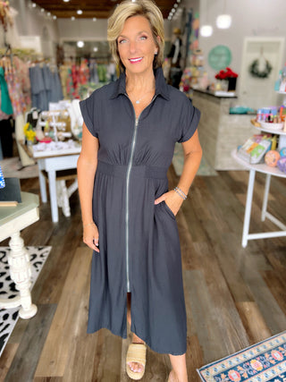 A woman modeling a dark gray midi shirt dress with a front zipper, short sleeves, and side pockets inside a boutique.