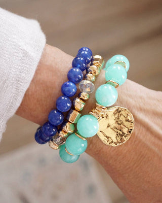 Wrist wearing multiple beaded bracelets with blue, green, and gold colors on a blurred background.
