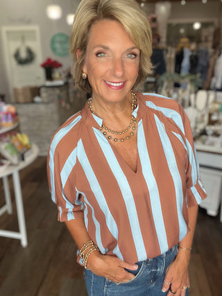 A smiling blonde woman wearing a brown and light blue vertically striped blouse with rolled sleeves, paired with blue jeans and layered gold necklaces, standing inside a boutique.