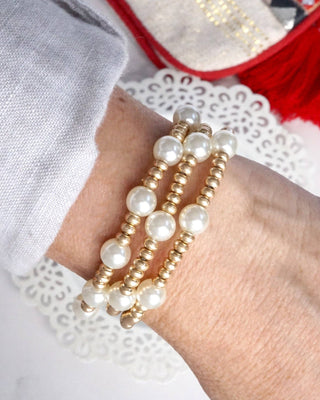 Close-up of a wrist wearing multiple pearl and gold bracelets with a red and white background.