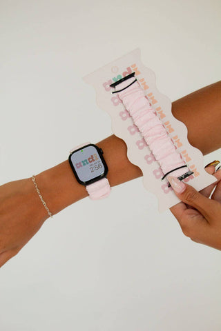 A person wearing a light pink smartwatch band with a black watch, holding a matching light pink fabric scrunchie on a decorative display card against a plain white background.