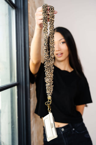 A woman in a black t-shirt holds up a leopard print fabric lanyard with a white card holder and gold metal clasp. The background shows a window frame and a neutral wall.