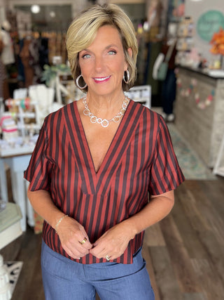A woman with blonde hair stands in a boutique wearing a rust and black vertically striped short-sleeve blouse with a V-neck, paired with blue jeans.