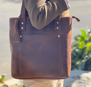 Brown leather tote bag with a hat on top, held by a person against a blurred natural background.