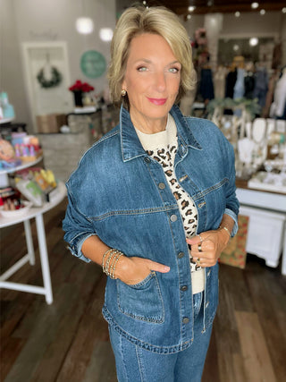 A woman with blonde hair wearing a blue denim jacket over a white top with a leopard print pattern, standing inside a boutique store.