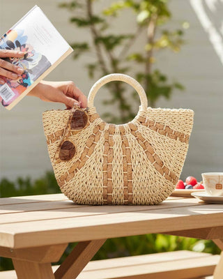 Woven handbag on a table with a cup and saucer, book, and potted plant in the background.