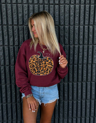 A woman wearing a burgundy crewneck sweatshirt with a large leopard-print pumpkin graphic on the front, paired with light blue denim shorts, standing against a dark gray textured wall.