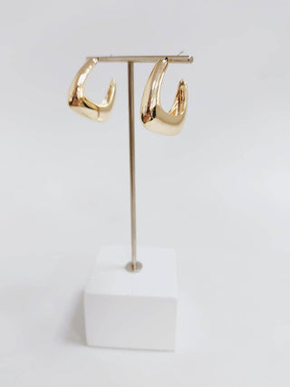 A pair of chunky, polished gold hoop earrings displayed on a minimalist gold T-stand with a white cube base against a clean white background.