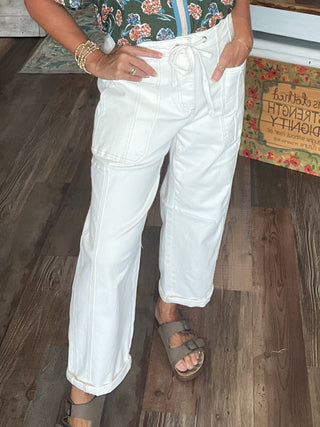 A person wearing white wide-leg cropped pants with a drawstring waist, paired with taupe double-strap sandals and a floral top, standing on a wood floor.