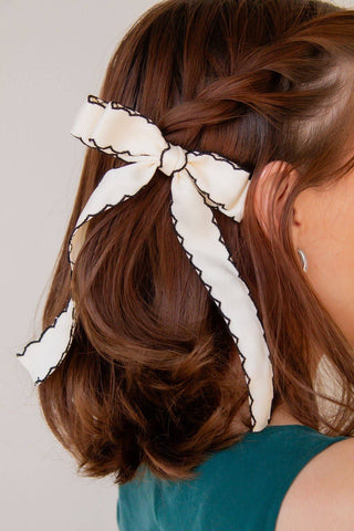 A cream-colored satin hair bow with black scalloped trim, styled in a side bun on a model with brown hair.