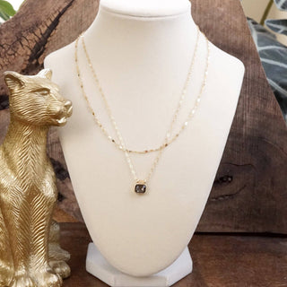 A double-strand gold necklace with a small square dark gemstone pendant, displayed on a white jewelry bust. A gold panther figurine sits to the left, and a rustic wooden background is visible behind the bust.
