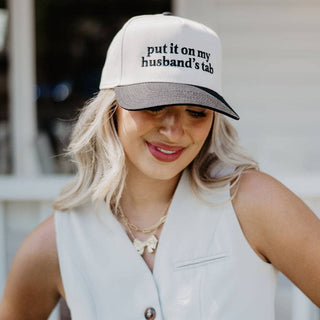 Woman wearing a cap with text 'put it on my husband's tab' outdoors.