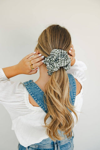 A woman shown from the back wearing a black and white gingham scrunchie in her long blonde-highlighted ponytail, styled with a white blouse and denim overalls against a light gray background.