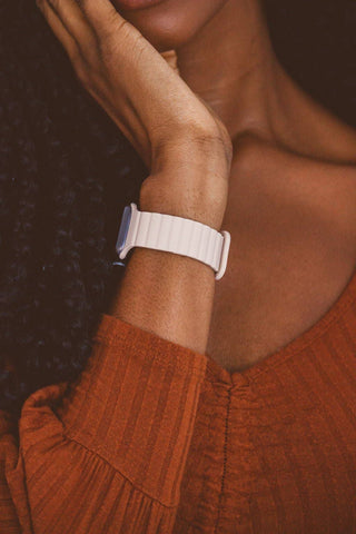 A close-up of a person wearing a nude silicone smartwatch band with a rectangular watch face, paired with a rust-orange ribbed knit top against a dark background.