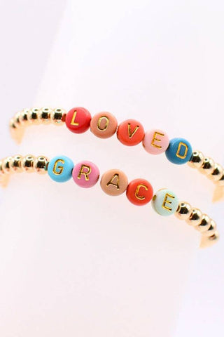 Two beaded stretch bracelets displayed on a white cylindrical stand. The top bracelet features gold-toned beads spelling 'LOVED' in red, orange, pink, and blue, with gold lettering.