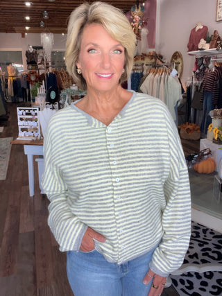 A woman with short blonde hair wearing a light gray and buttercream striped button-up cardigan sweater and light blue jeans, standing inside a clothing boutique.