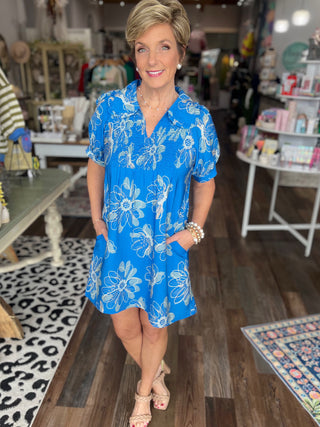 Woman in a blue floral dress standing in a store with various items on display.
