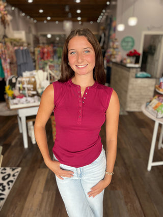 A woman smiling in a boutique shop, wearing a sleeveless henley top in a vibrant magenta pink and light wash jeans.