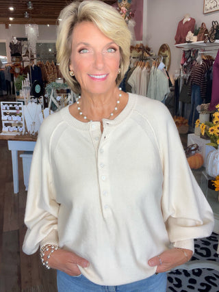 A smiling blonde woman wearing a cream-colored knit henley sweater with button-down front and balloon sleeves, standing inside a boutique store.