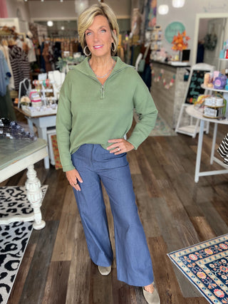 A woman stands in a retail boutique wearing a sage green half-zip pullover sweater and blue wide-leg pants. The store has wood floors, clothing racks, and display tables in the background.