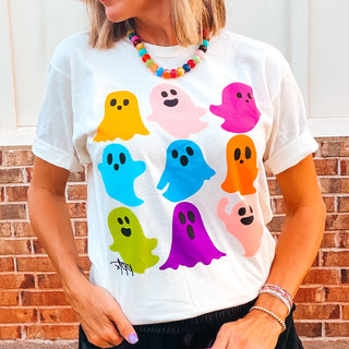 A woman wearing a white graphic t-shirt with a colorful print of cartoon ghosts in yellow, pink, blue, orange, green, purple, and light pink, posed against a brick wall background.