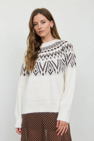 A woman modeling a cream knit sweater with a dark brown geometric Fair Isle pattern across the yoke, paired with brown polka-dot pants, against a plain light gray background.
