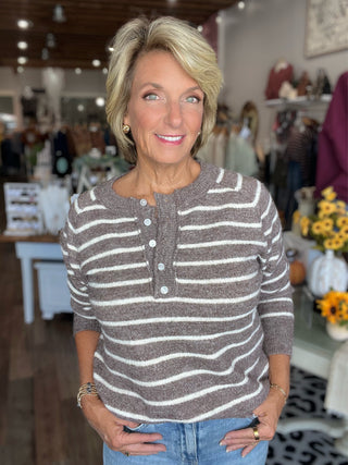 A smiling blonde woman wearing a brown and cream horizontally striped henley sweater, standing in a boutique store.