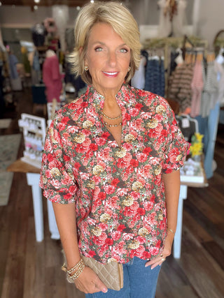 A woman with blonde hair stands in a clothing store wearing a short-sleeve button-up blouse with a red, pink, and cream floral print on a dark background, paired with blue jeans and holding a quilted tan clutch.