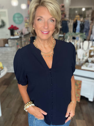 A woman with short blonde hair wearing a navy blue short-sleeve button-up blouse and blue jeans, standing in a boutique setting.